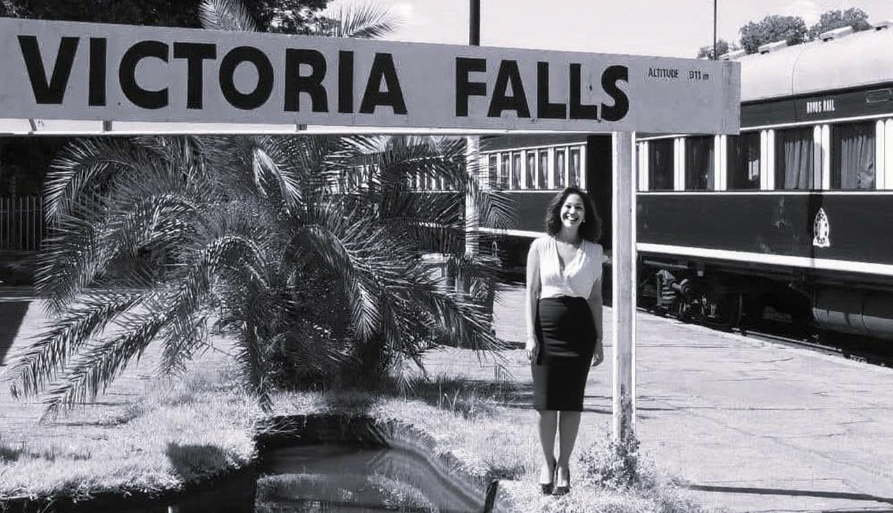 Lady standing under the sign for Victoria Falls Train Station, with the Rovos Rail train having stopped alongside